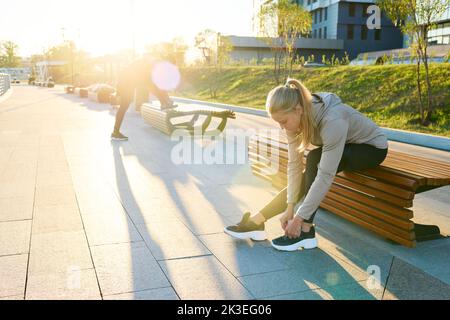 Young blond sportswoman in activewear sitting on bench in urban environment and bending over sneaker while tying shoelace Stock Photo