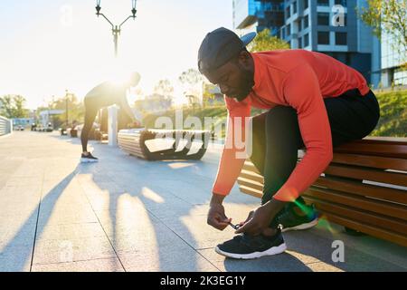 Young African American sportsman in red sport jacket and black leggins tying shoelace of sneaker while bending forwards over leg Stock Photo