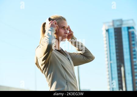 Side view of young blond woman in grey sport jacket putting headphones on her head while standing against blue sky and skyscraper Stock Photo