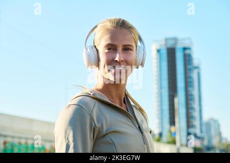 Happy young blond woman in white leather headphones and grey sport jacket looking at camera in urban environment after workout Stock Photo