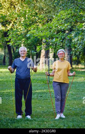 Senior spouses wear sportswear doing exercises outdoor in summer park in the morning using resistance rubber bands. Healthy lifestyle, active retired Stock Photo