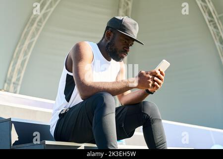 Young serious black man in activewear texting in smartphone on sunny morning while having rest after jogging or sports training Stock Photo