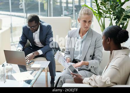 Young confident blond female economist showing document with financial data to African American colleague with tablet Stock Photo