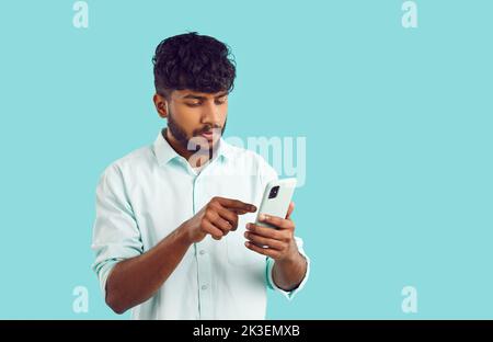 Young focused Indian man holding mobile phone reading latest news stands on turquoise background Stock Photo