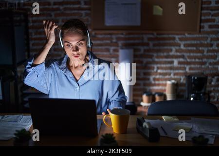 Beautiful brunette woman working at the office at night angry and mad raising fist frustrated and furious while shouting with anger. rage and aggressi Stock Photo