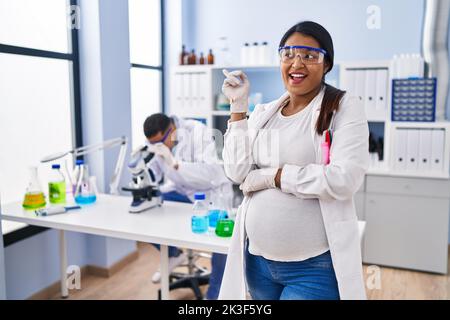 Young hispanic woman expecting a baby working at scientist laboratory smiling happy pointing with hand and finger to the side Stock Photo