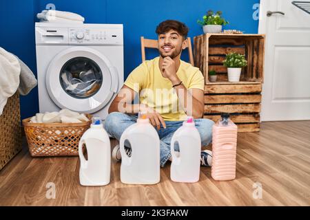 Arab man with beard doing laundry sitting on the floor with detergent bottle smiling looking confident at the camera with crossed arms and hand on chi Stock Photo