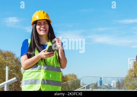 young latin venezuelan engineer woman, wearing yellow helmet and vest standing outdoors laughing out loud because of a joke texted to her phone, techn Stock Photo