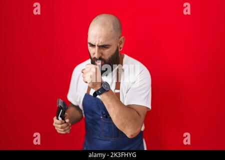Young hispanic man with beard and tattoos wearing barber apron holding razor feeling unwell and coughing as symptom for cold or bronchitis. health car Stock Photo