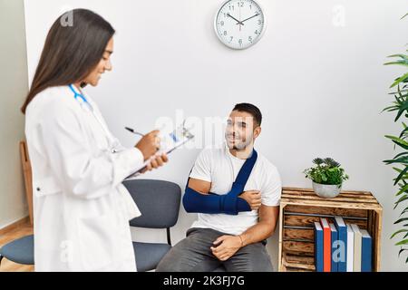 Youg latin injuried man wearing sling talking with the doctor at clinic waiting room. Stock Photo