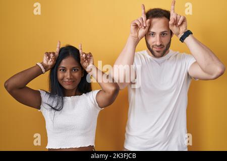 Interracial couple standing over yellow background doing funny gesture with finger over head as bull horns Stock Photo