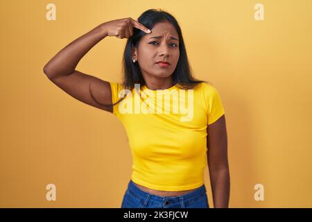Young indian woman standing over yellow background pointing unhappy to pimple on forehead, ugly infection of blackhead. acne and skin problem Stock Photo