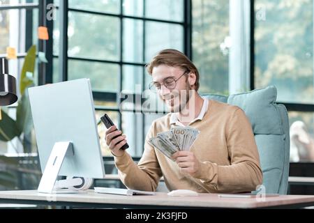 Young successful businessman working inside modern loft office, happy man looking at phone screen, reading profit news, holding piles of cash. Stock Photo
