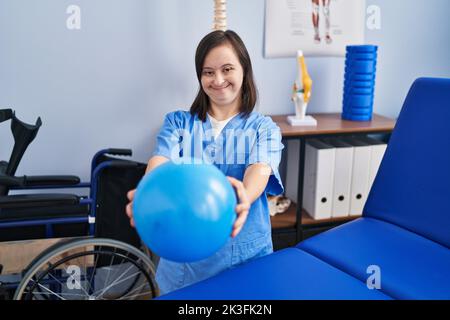 Down syndrome woman wearing physiotherapy uniform holding ball at physiotherapist clinic Stock Photo