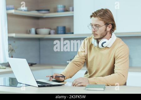 Young serious and focused man studying remotely at home, student in kitchen wearing glasses using laptop for distance learning taking notes on study material. Stock Photo