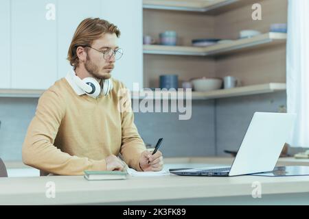 Young serious and focused man studying remotely at home, student in kitchen wearing glasses using laptop for distance learning taking notes on study material. Stock Photo