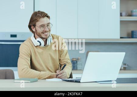 Young male entrepreneur working at home on paper work, sitting in the kitchen at the table, using a laptop for remote work as a freelancer. Stock Photo