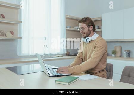 Young successful and happy businessman working from home, man using laptop for remote work, freelancer smiling in casual clothes sitting in kitchen at table. Stock Photo