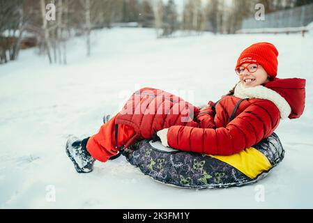 a teenage girl in a red jacket in a snowy forest rides in a tubing on a cold day. Stock Photo