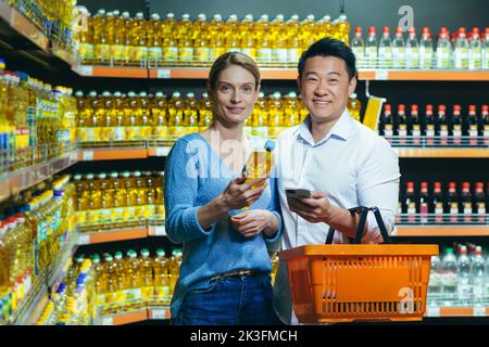 A young international couple, a woman and an Asian man, are choosing oil in a supermarket. They are holding a bottle of oil, a basket and a phone. They are looking at the camera and smiling. Stock Photo