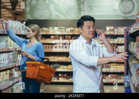 A young Asian man is shopping in a supermarket. Stands near the shelf with goods, chooses, talks on the phone, consults. Shoppers in the background. Stock Photo