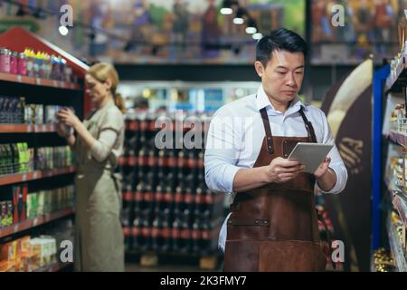 Portrait of young handsome Asian man, shop owner, supermarket worker. Standing in an apron near the shelves with goods, writing in a notebook. Female employee in the background. Stock Photo