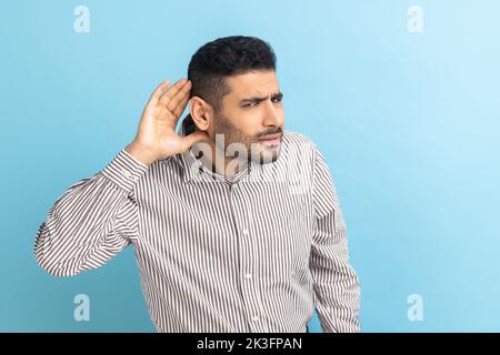 Concentrated nosy businessman with beard holding hand near ear, listening to interesting talks and private secrets, spying, wearing striped shirt. Indoor studio shot isolated on blue background. Stock Photo