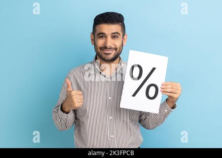 Smiling smart businessman showing paper with percent sign inscription, looking at camera, showing thumb up, wearing striped shirt. Indoor studio shot isolated on blue background. Stock Photo