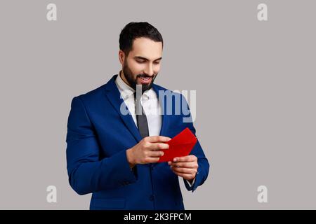 Handsome bearded businessman pulling letter from red envelope, holding greeting card and smiling joyfully, wearing official style suit. Indoor studio shot isolated on gray background. Stock Photo