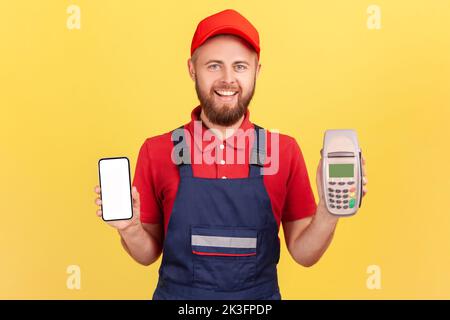 Portrait of smiling delighted bearded worker man standing and holding pos terminal and smartphone with blank screen for your advertisement. Indoor studio shot isolated on yellow background. Stock Photo