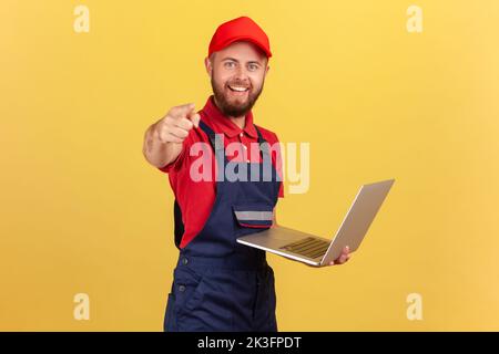 Delighted worker man wearing blue overalls, red T-shirt and cap working on laptop, pointing finger at camera, showing for you a new online service. Indoor studio shot isolated on yellow background. Stock Photo