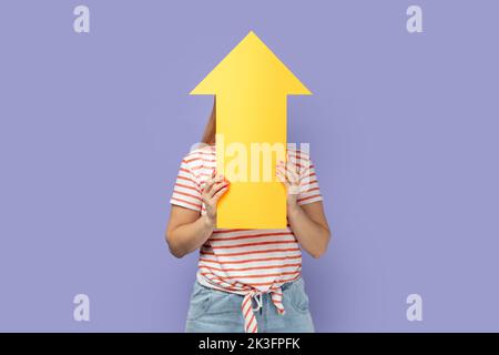 Portrait of anonymous unknown blond woman wearing striped T-shirt holding big yellow arrow pointing up, showing increase in career. Indoor studio shot isolated on purple background. Stock Photo