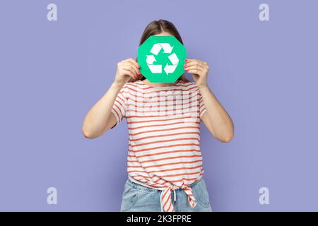 Portrait of unknown anonymous woman wearing striped T-shirt holding green recycling sign and hiding her face behind ecology symbol. Indoor studio shot isolated on purple background. Stock Photo