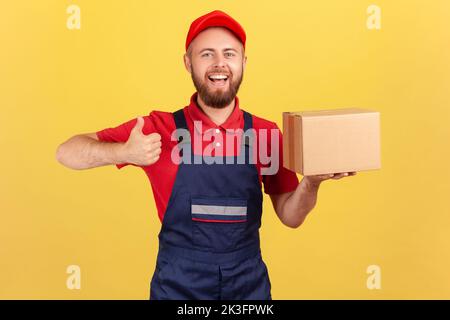 Cargo transportation, delivery service. Smiling courier man in uniform holding cardboard box and showing thumb up, delivering parcel to client. Indoor studio shot isolated on yellow background. Stock Photo