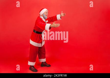 Full length profile shot of elderly man with gray beard wearing santa claus costume reaching hands out, stretching arms to hug someone. Indoor studio shot isolated on red background. Stock Photo