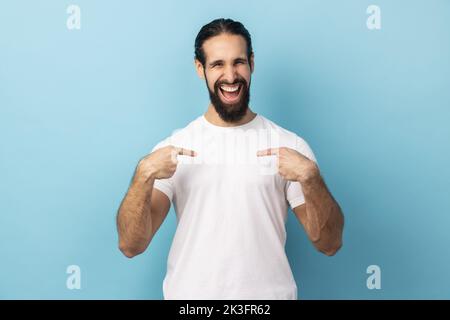 Portrait of delighted man with beard wearing white T-shirt pointing fingers on himself and looking at camera with toothy smile, extremely satisfied. Indoor studio shot isolated on blue background. Stock Photo