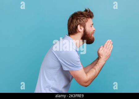 Please, I'm begging. Side view of bearded man holding arms in prayer, asking help or forgiveness with imploring eyes, sincere asking permission. Indoor studio shot isolated on blue background. Stock Photo