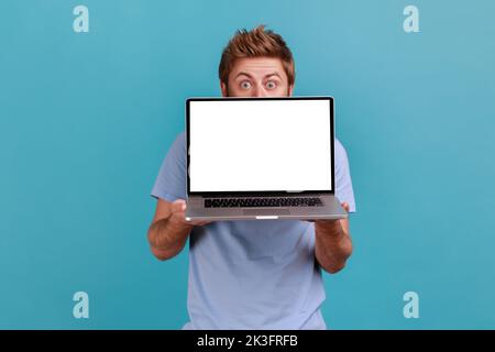Portrait of man hiding behind white empty screen laptop, surprised with advertising area, looking at camera with big eyes, updating operation system. Indoor studio shot isolated on blue background. Stock Photo