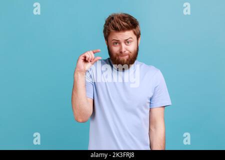 Portrait of disappointed bearded man showing a little bit gesture, dissatisfied with low rating, measuring scale, frowning face and looking at camera. Indoor studio shot isolated on blue background. Stock Photo