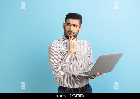 Portrait thoughtful pensive man with beard holding laptop and holding chin looking away, thinking over new business project, wearing striped shirt. Indoor studio shot isolated on blue background. Stock Photo