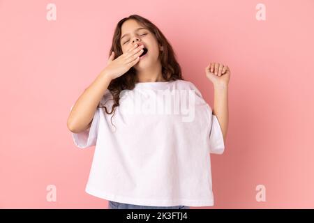 Portrait of adorable sleepy little girl wearing white T-shirt standing and yawning with closed eyes and raised arm, covering mouth with palm. Indoor studio shot isolated on pink background. Stock Photo