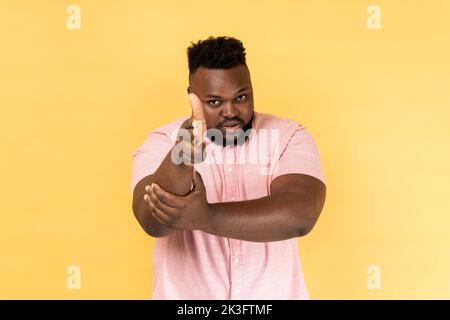 I'll kill you. Portrait of man wearing pink shirt pointing finger guns to camera and looking aggressive, threatening to shoot, hands imitating weapon. Indoor studio shot isolated on yellow background. Stock Photo