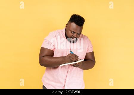 Portrait of astonished man wearing pink shirt writing in notebook, completing to-do list or studying, having surprised expression. Indoor studio shot isolated on yellow background. Stock Photo