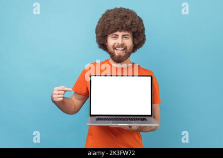 Portrait of delighted man with Afro hairstyle wearing orange T-shirt standing showing empty display of laptop, pointing at screen, advertising area. Indoor studio shot isolated on blue background. Stock Photo