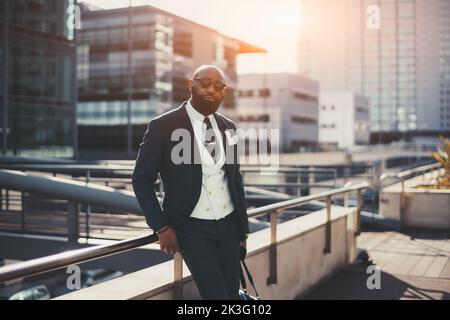 A confident mature bald bearded African man entrepreneur in sunglasses and a tailored suit is leaning against railing while standing on the street of Stock Photo