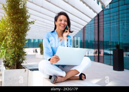 Businesswoman use laptop outdoor sitting near office building. Caucasian female business person 30 years have phone conversation and smile. Remote work freelancer concept. Freelance job its new normal Stock Photo