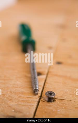 Green screwdriver with square tip and metal screw head sticking out from plywood floor surface inside home. Stock Photo