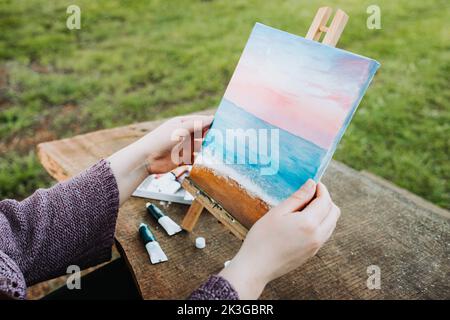 Unrecognizable woman's hands holding her own beach scenery oil painting. Amateur painter. Stock Photo
