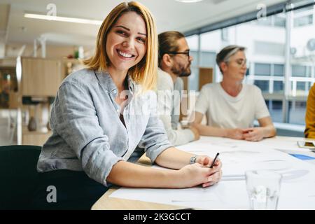 Cheerful young designer smiling at the camera while sitting in a meeting with her colleagues. Team of innovative businesspeople sharing creative ideas Stock Photo