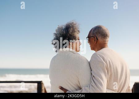 Mature lovebirds by the ocean. Rearview of a romantic senior couple having a cheerful chat on a seaside foot bridge. Retired elderly couple enjoying s Stock Photo